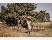 Grüne Wiese mit einem Hochzeitsbogen aus Holz, dekoriert mit Blumen und Stoff, umgeben von Bäumen und hohen Gräsern. Vor dem Bogen befinden sich Reihen von Holzstühlen mit weißen Kissen, die eine Sitzordnung für eine Zeremonie bilden.