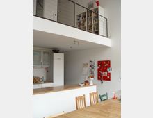 Wohnbereich mit offener Küche im Vordergrund und einem darüberliegenden Hochgeschoss. Die Küche verfügt über Holzarbeitsplatten, helle Schränke und eine Spüle. Im Hintergrund ist ein Esstisch mit Holzstühlen sowie eine rote Pinnwand an der Wand zu sehen.