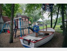 Spielplatz mit Piratenthema, bestehend aus einem Holzboot mit einem Steuerrad und einer Piratenflagge, umgeben von Rindenmulch und Bäumen. Im Hintergrund stehen ein Klettergerüst mit Rutsche und ein Pavillon.