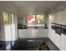 Moderne, helle Küche mit weißen, griffleistenlosen Schränken, schwarzen Arbeitsflächen und integrierten Backöfen an einer Wand mit grauer Steinoptik. Große Fenster und eine Glastür lassen viel Tageslicht herein; im Vordergrund steht eine Kücheninsel mit schwarzer Platte.