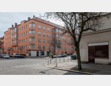 Straßenszene mit einem mehrstöckigen Wohngebäude in einem orangefarbenen Ton mit weißen Fensterrahmen. Im Vordergrund verlaufen eine Kreuzung, ein Gehweg mit Pollern und ein Baum ohne Laub.