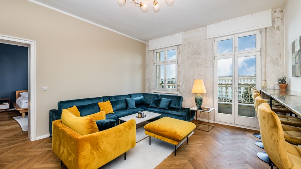 Helles Wohnzimmer mit Fischgrätparkett, großem Ecksofa in Dunkelblau sowie gelbem Sessel und Hocker um einen niedrigen Couchtisch auf Teppich. Hohe Fenster und eine Balkontür mit Blick nach draußen, dazu eine Barzeile mit gepolsterten Hockern an der rechten Wand und eine Deckenleuchte.