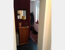 Flur in einer Wohnung mit dunklem Fliesenboden und einem roten Teppich. An der roten Wand steht eine hölzerne Kommode mit einem Spiegel darüber, daneben eine Garderobe mit Kleidung und ein Fenster auf der rechten Seite.