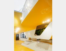 Innenansicht eines modernen Gebäudes mit gelben Böden und Decken sowie weißen, geschwungenen architektonischen Elementen. Eine Wandöffnung integriert eine grüne Pflanzenwand, und große Fenster lassen Tageslicht herein.