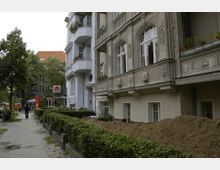 Straßenszene mit mehrstöckigen Wohnhäusern im Altbaustil, verziert mit Ornamenten und Balkonen; vor einem Gebäude ist Erdreich aufgeschüttet, daneben verläuft ein Bürgersteig mit Hecken und Bäumen. Im Hintergrund sind weitere Gebäude und eine Ampel zu sehen.