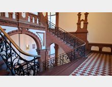 Innenansicht eines Treppenhauses mit roten Ziegelbögen und dekorativem Mauerwerk, einer geschwungenen Treppe und einem schwarzen schmiedeeisernen Geländer. Der Boden ist mit rot-weißen Schachbrettfliesen ausgelegt, an den Wänden hängen gerahmte Bilder.