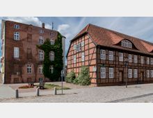 Backsteingebäude und Fachwerkhaus in einer gepflasterten Platzumgebung. Das linke Gebäude ist ein simples Ziegelbauwerk, teilweise von Efeu bewachsen, während das rechte ein klassisches Fachwerkhaus mit roten Ziegeln und weißen Fensterrahmen sowie einem geschwungenen Dach ist.