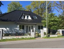 Einfamilienhaus mit weißer Fassade und schwarzem Satteldach, umgeben von einem Metallzaun und einem Vorgarten mit blühenden Tulpen. Im Hintergrund sind Bäume und ein klarer blauer Himmel zu sehen.