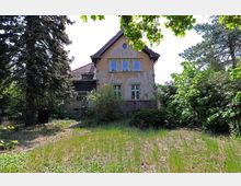 Ein älteres, zweigeschossiges Haus mit verwitterter Fassade und rotem Ziegeldach, umgeben von dichter Vegetation, darunter Laub- und Nadelbäume sowie Sträucher. Im Vordergrund liegt ein ungepflegter Garten mit Gras und vereinzeltem Bewuchs.