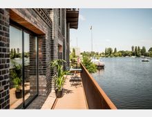 Backsteinfassade eines Gebäudes mit einem schmalen Balkon, der Blick auf einen Fluss bietet. Im Hintergrund sind Boote, Bäume und ein Turm zu sehen, während auf dem Balkon Topfpflanzen und ein kleiner Tisch mit Stühlen stehen.