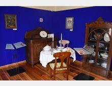 Ein historischer Raum mit einem Tisch, der mit weißer Tischdecke, Kerzenhaltern, Geschirr und Backwaren gedeckt ist. Im Hintergrund sind ein Holzsideboard, eine Vitrine mit Porzellan und ein tiefblauer Anstrich an den Wänden zu sehen, ergänzt durch kleine Bilder und Informationstafeln.