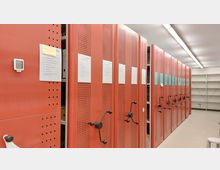 Rote rollbare Archivregale mit perforierten Metalltüren und Handkurbeln in einem gut beleuchteten Lagerraum. Auf den Regalen sind Papieretiketten angebracht, daneben befinden sich leere weiße Regale an der Wand.