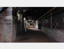 Ein langer, dunkler Korridor in einem verlassenen Industriegebäude mit sichtbaren Rohren an der Decke, brüchigen Betonwänden und teilweise zerstörten Glasbausteinfenstern. Eine Leiter lehnt an der linken Wand, und der Boden ist mit Staub und Schmutz bedeckt.