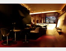 Dunkel gestaltete Lounge-Bar mit gedämpfter Beleuchtung, modernen schwarzen Sitzmöbeln und kleinen runden Tischen. Im Hintergrund befindet sich eine beleuchtete Theke mit Barhockern und einer großen Regalwand voller Flaschen.
