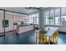 Moderne Küche mit heller Einbauküchenzeile in Pastellfarben, zentraler Kücheninsel und großem Esstisch mit transparenten Stühlen. Große Fenster lassen viel Tageslicht herein, und die weiß gestrichenen Wände sowie die Sicht auf ein benachbartes Gebäude mit roten Ziegeldächern setzen einen hellen Akzent.