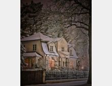 Verschneites zweistöckiges Wohnhaus mit steilem Satteldach und mehreren Gaubenfenstern, vor einem Zaun und winterlichen Sträuchern. Große, schneebedeckte Baumäste rahmen die Szene ein, die Fenster leuchten warm in einer nächtlichen Umgebung.