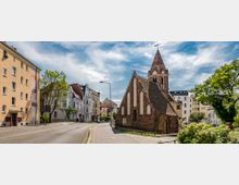Straßenszene mit einer kleinen, historischen Kirche aus Backstein mit spitzem Turm in der Bildmitte, umgeben von mehrstöckigen Wohnhäusern und Bäumen. Der Himmel ist leicht bewölkt, und die Straße verläuft geschwungen entlang der Gebäude.