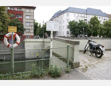 Szene an einem Kanal mit einer Metallabsperrung, an der ein Rettungsring befestigt ist. Im Hintergrund sind eine Brücke, ein geparktes Motorrad sowie Wohngebäude mit Bäumen zu sehen.