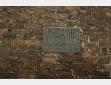 Eine Ziegelmauer mit einer eingelassenen Tafel aus Stein, die auf einen Rest der alten Stadtmauer hinweist. Der Text beschreibt die Stadtbefestigung aus der ersten Hälfte des 14. Jahrhunderts.