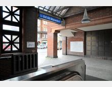 U-Bahnhof Alt-Mariendorf in Berlin, mit roten Backsteinwänden, einer blauen Stationsbeschilderung und einer Rolltreppe im Vordergrund. Im Hintergrund ist die Straße mit Gebäuden und geparkten Autos zu sehen.