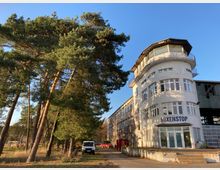 Ehemaliges funktionales Gebäude mit weißer Fassade und Rundbau an der Vorderseite, auf dem Schriftzug „Boxenstop“ steht, daneben Kiefern und geparkte Fahrzeuge auf einer asphaltierten Fläche. Der Himmel ist klar und blau.