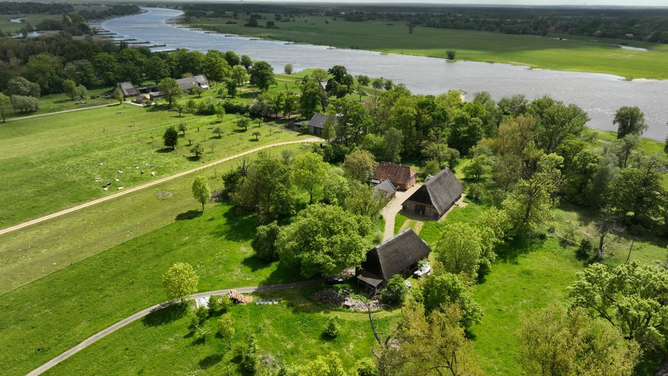 Luftaufnahme einer ländlichen Flusslandschaft mit breitem, geschwungenem Flusslauf, grünen Wiesen und Baumgruppen. Im Vordergrund liegen mehrere Bauernhofgebäude mit teils reetgedeckten Dächern an einem Feldweg, dahinter erstrecken sich offene Felder bis zum Horizont.