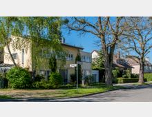 Wohnstraße mit freistehenden Häusern im Stil moderner Einfamilienhäuser, umgeben von Bäumen und gepflegten Gärten. Im Vordergrund stehen Straßenschilder mit den Bezeichnungen „Ravenweg“ und „Kastanienweg“.