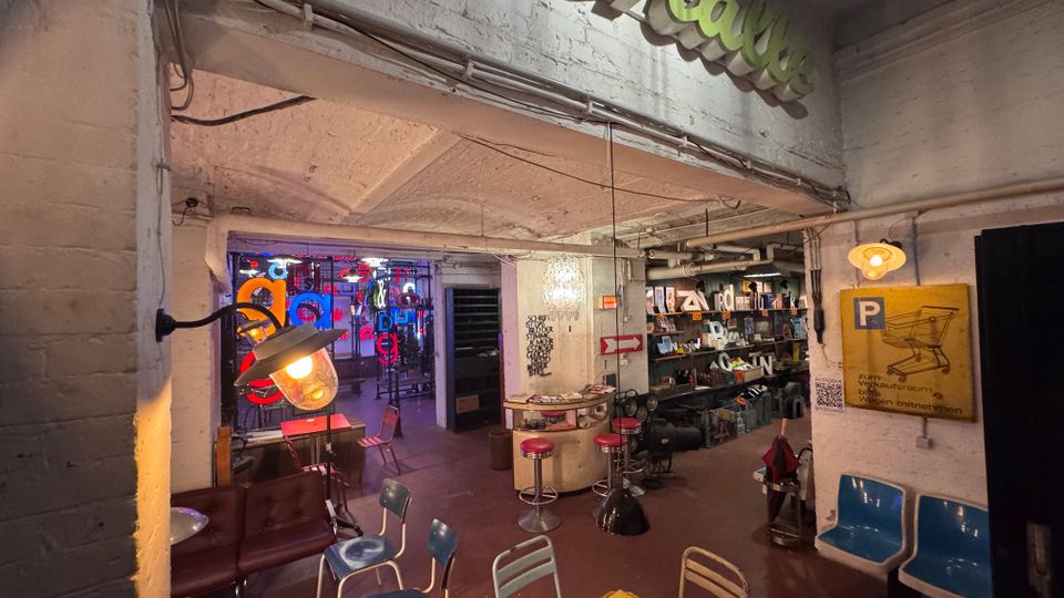 Ein industriell gestalteter Innenraum mit weißen Ziegelwänden, freiliegenden Rohren und einer Mischung aus Retro-Möbeln wie bunten Stühlen und Neonlichtern. Im Hintergrund befinden sich eine Bar mit Hockern, eine Bücherwand und bunte Leuchtschrift-Dekorationen.