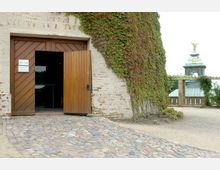 Ein Backsteingebäude mit einer teilweise geöffneten hölzernen Eingangstür und efeubewachsenen Wänden, davor ein gepflasterter Weg; im Hintergrund ist ein kleiner Turm mit einem Dach aus Kupfer und einer goldenen Figur sichtbar.