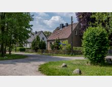 Straßenszene in einem Wohngebiet mit einzelnen Einfamilienhäusern, die Ziegeldächer und Vorgärten haben. Umgeben von Grünflächen, Bäumen und einem unbefestigten Weg.