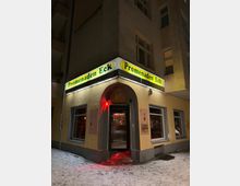 Eckeingang eines Mehrfamilienhauses bei Nacht mit beleuchtetem Eck-Schild „Promenaden Eck“ über einem Rundbogenportal und mehreren Fenstern. Vor dem Eingang liegt ein schmaler, verschneiter Gehweg; rotes Licht beleuchtet den Bereich um die Tür.