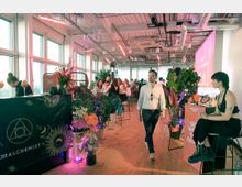 Bright modern event space with large floor-to-ceiling windows, wooden floors, and an exposed industrial ceiling with ducts and hanging lights. Plants and vendor-style counters line the room, with bar stools and a pink-lit projection wall at the far end.