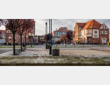 Platz mit gepflastertem Weg, umgeben von Bäumen mit herbstlichem Laub, im Hintergrund Gebäude aus rotem Backstein und moderner Glasfassade; rechts ein historisches Gebäude mit der Aufschrift "Konrad".