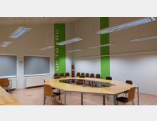 Ein moderner Seminarraum mit hellen Holztischen, die in einer offenen U-Form angeordnet sind, umgeben von Stühlen mit Holzrahmen und gepolsterten Sitzflächen. Die Wände sind teilweise grün gestaltet, und an einer Wand steht in großen weißen Buchstaben „UP TO YOU“.