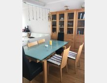 Esszimmer mit hellem Holzboden, einem Glastisch und einer Kombination aus Holzstühlen mit gestreiften Sitzkissen und schwarzen Lederstühlen. Im Hintergrund stehen ein weißes Sofa und ein großer Holzvitrinenschrank mit Glastüren, gefüllt mit Geschirr und Büchern.