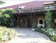 Hof mit gepflastertem Innenhof, umgeben von einem Backsteingebäude mit weißen Türen und kleinen Fenstern; ein Balkon mit Holzgeländer und Blumenkästen läuft entlang der oberen Etage. Im Vordergrund befinden sich blühende Rosenbüsche und Rankenpflanzen an den Wänden.