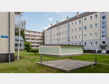 Mehrgeschossige Wohngebäude in einem urbanen Wohngebiet mit verputzten Fassaden in beige und grauen Tönen. Im Vordergrund eine grüne Rasenfläche mit einem flachen Nebengebäude und einem betonierten Bereich.