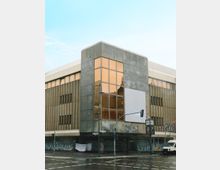 Eckgebäude aus den 1970er Jahren mit einer Fassade aus Beton und Glas, teilweise mit Graffiti bedeckt. Im Erdgeschoss sind Schaufenster und Eingänge, umgeben von einer nassen Straße mit Straßenlaternen und Verkehrsampeln; links ein Werbebanner und rechts ein Lieferwagen.