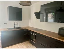 Moderne L-förmige Küche mit mattschwarzen Schränken und Holzarbeitsplatte, integriertem Spülbecken mit schwarzer Armatur sowie Herd und Backofen. An der Wand befinden sich glänzende, schwarze Hängeschränke und eine schräg montierte Dunstabzugshaube; heller Holzboden und Deckenleuchte ergänzen den Raum.