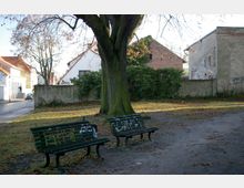 Ein kleiner, baumbestandener Platz mit zwei mit Graffiti besprühten Bänken im Vordergrund. Im Hintergrund sind eine Mauer, Sträucher sowie Gebäude mit Ziegeldächern und Putzfassaden zu sehen.