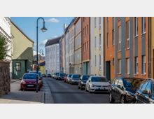 Straßenszene mit farbenfrohen Wohnhäusern in unterschiedlichen Pastelltönen, darunter Orange, Blau und Gelb. Entlang der gepflasterten Straße sind parkende Autos auf beiden Seiten sichtbar, und Laternen im klassischen Stil säumen den Gehweg.