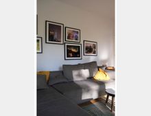 Wohnzimmer mit einer großen grauen Eckcouch, dekoriert mit gelben Kissen. An der weißen Wand darüber hängen mehrere gerahmte Bilder von urbanen Landschaften und Architektur, die in einer Rasteranordnung platziert sind.