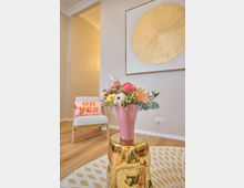 Modernes Wohnzimmer mit einem hellen Holzfußboden und neutralen Wandfarben. Im Vordergrund ein goldener Hocker mit einer rosafarbenen Vase mit bunten Blumen, im Hintergrund ein Sessel mit einem rosa Kissen und ein großes, rundes, goldenes Wandbild.