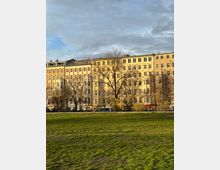 Wohnhäuser in hellen Pastelltönen mit mehreren Stockwerken und zahlreichen Fenstern, umgeben von kahlen Bäumen. Im Vordergrund befindet sich eine weitläufige, grüne Rasenfläche unter einem leicht bewölkten Himmel.