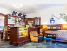 Gemütliches Café mit Holztheke, Glasvitrine, Getränkeregalen und Kreidetafeln an der Wand. Im Sitzbereich stehen ein beiger Sessel, ein gepolstertes Sofa, ein Bücherregal und eine Retro-Spielmaschine, ergänzt durch warme Beleuchtung und Dekoration.