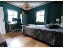 Schlafzimmer mit dunkelgrünen Wänden, zwei Fenstern mit passenden Vorhängen und Holzboden in Fischgrätenmuster. Das Zimmer enthält ein Bett mit grauer Tagesdecke, einen Schreibtisch mit Bürostuhl und Computer sowie einen Wandschrank mit einer Gitarre darauf.