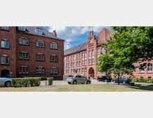 Backstein-Gebäudekomplex mit historischen Elementen und Satteldächern, umgeben von parkenden Autos und einer Grünfläche mit Bäumen; im Hintergrund ist ein Durchgangsbogen in einem der Gebäude sichtbar.