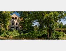 Ehemaliges, verlassenes Gebäude mit beschädigter Fassade und teilweise zerstörten Fenstern, von dichter Vegetation und Bäumen umgeben. Links das zweistöckige Hauptgebäude, rechts ein flaches, unscheinbares Nebengebäude.