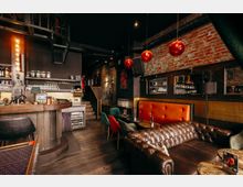 Innenansicht einer Bar im Industrial-Stil mit Backsteinwand, dunkler Decke und schwarzer Metall-Spindeltreppe. Zu sehen sind eine Holztheke mit Flaschenregalen, rote Kugel-Pendelleuchten sowie Lounge-Sitzplätze mit Chesterfield-Ledersofa und kleinen Tischen.