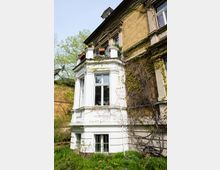 Historisches Wohngebäude mit einer Fassade aus gelbem Klinker und weiß verputztem Erker. Der Erker verfügt über große Fenster, daneben ranken Kletterpflanzen an der Wand, und im Vordergrund befinden sich ein gepflegter Garten mit Blumen und Sträuchern.